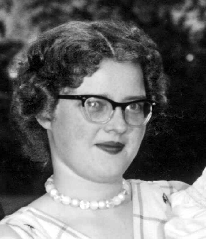 Judy Taylor black and white photo with short wavy black hair. Cat eyeglasses with black upper and clear on lower part of frames. Choker. Red lipstick. Not smilin.