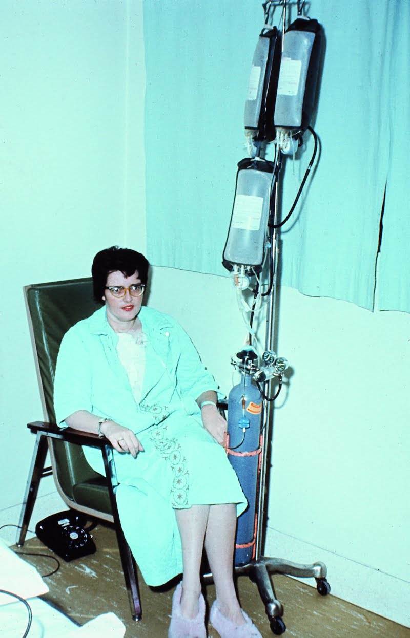 Judy Taylor in Toronto General Hospital sitting in a black chair in blue dressing gown with IV pole, air tank at the bottom and thick IV type bag hanging from top. Tubes snaking into her.