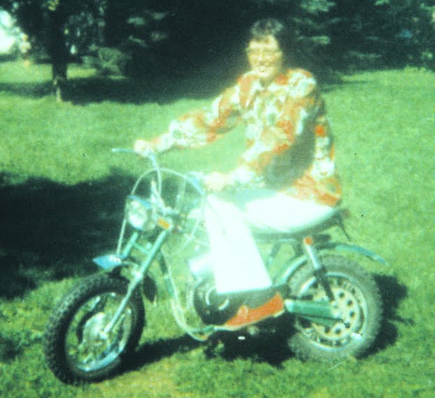Judy Taylor posing on a small motorcycle on a lawn. Garish 1970s shirt and white pants. Short straight black hair.