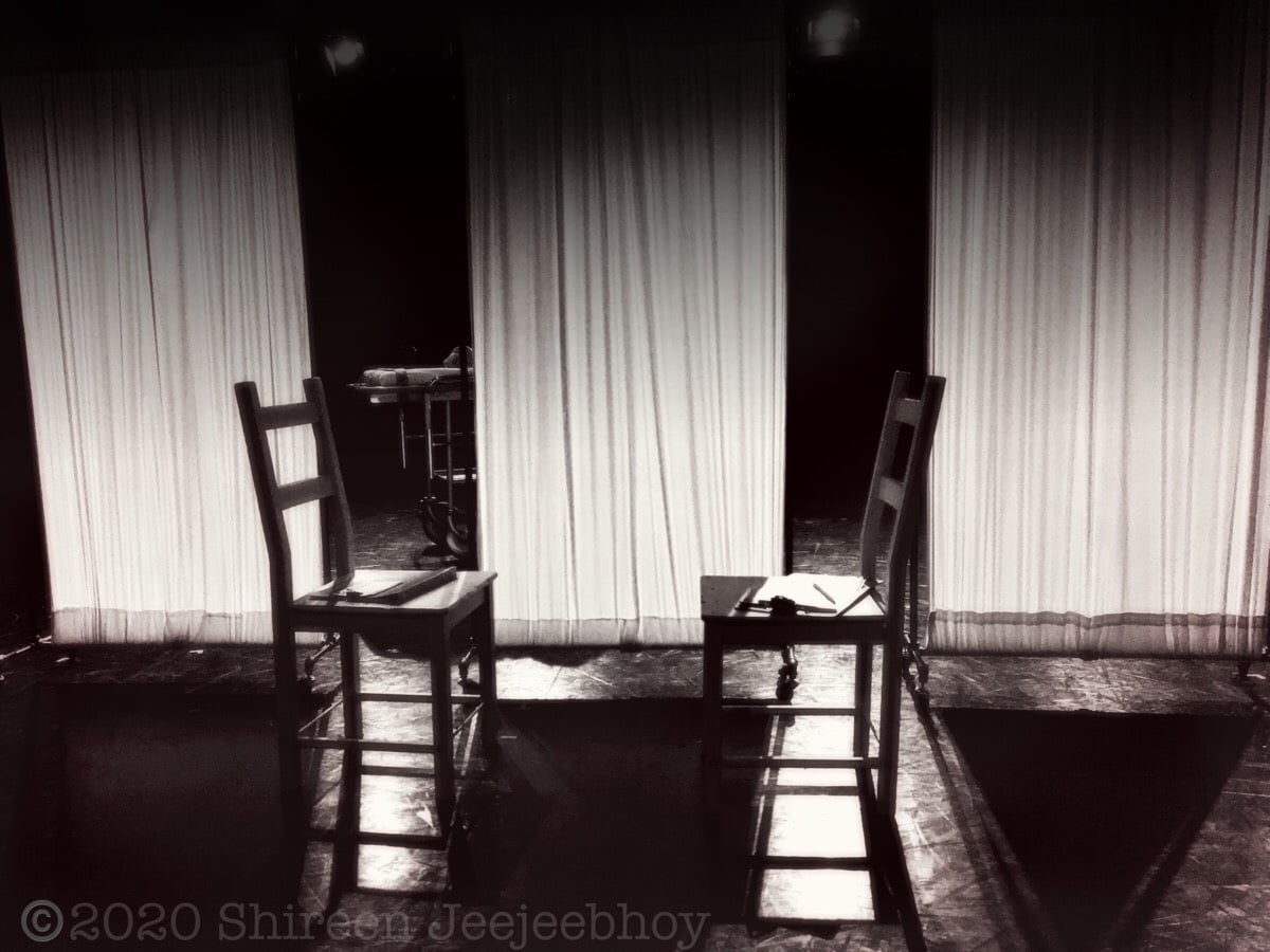 Two empty ladderback chairs facing each other on an empty stage with last-century white surgical curtain panels behind. They're spotlighted.
