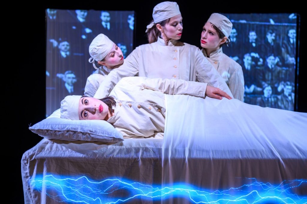 The two Penfields and Shapiro standing behind Mrs Gold on the operating table as an audience projection watches. An opportunity to capture brain functions through electrical stimulation. Production photo by Dahlia Katz.