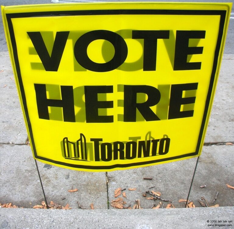 Plastic yellow sign with black letters Vote Here Toronto with city logo. On metal stilts planted in sidewalk. A few dead leaves on concrete.