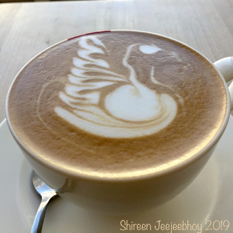 A cup of cappuccino, with white swan latte art in a wite cup, a tiny spoon partly hidden by the cup in lower left. On a wood table.