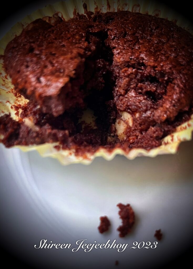 Close up of brownie in cupcake cup with a wedge cut out and a spotlight lighting up the front edge with a couple of rich dark brown crumbs. 