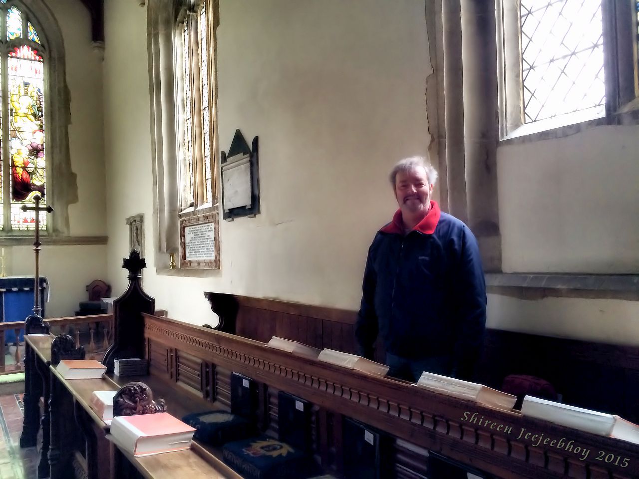 A man standing in a choir pew near the wall in his ancient stone church. A leaded glass window behind him with light streaming in from behind and the side. In the far left distance part of the stained glass window is visible.
